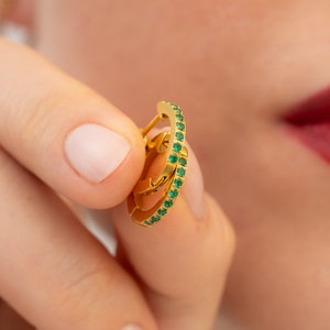 May include: Gold hoop earrings with a row of small green gemstones around the edge.