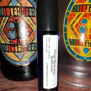 May include: A small, dark blue glass bottle with a white label that reads "Wizard Apothecary Butterbeer" and "pre diluted essential oil roll on 10ml". The bottle is sitting on a wooden surface.