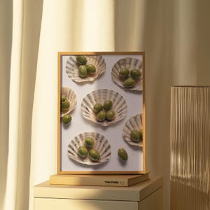Könnte beinhalten: Ein gerahmter Druck mit grünen Oliven, die in Jakobsmuschelschalen angeordnet sind. Der Druck hat einen Holzrahmen und liegt auf einem Stapel Bücher. Der Text "TOM FORD" ist auf dem Buchrücken zu sehen. Die Gesamtästhetik ist minimalistisch und elegant.
