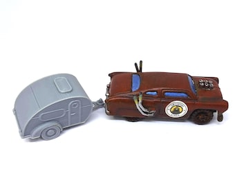 May include: A miniature brown car with a gray camper trailer attached. The car has a weathered appearance with blue window accents and a circular emblem. The trailer is gray with a rounded shape and a small window.