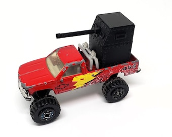 May include: A red toy truck with large black tires and a black box-like structure on the bed. The truck has a yellow and red design on the side and the number "454" is visible. The truck has a silver grill and headlights.