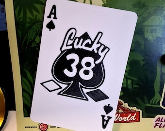 May include: A white playing card featuring the Ace of Spades with the words "Lucky 38" and the number "38" in black. The card is part of a set with a "Nuka-World" logo visible.
