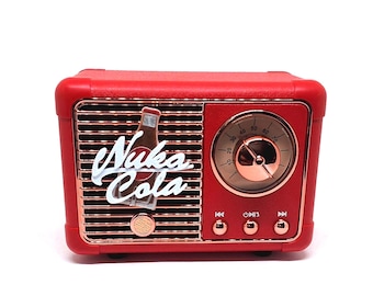 Pode incluir: Um alto-falante portátil vermelho com design retrô, apresentando uma grade de alto-falante e um dial de sintonia circular em ouro rosa. A frente exibe o logotipo Nuka-Cola e um gráfico de garrafa em branco. Três botões de controle em ouro rosa.