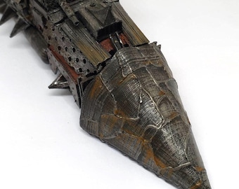May include: A detailed, metallic model of a war machine with a spiked front and a textured, weathered appearance. The model features a combination of dark gray, brown, and rust-colored accents, suggesting a post-apocalyptic or fantasy theme. The design includes various geometric shapes and perforations.