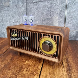 May include: A vintage-style radio with a wood-grain finish. The radio features a gold-colored grill, a tuning dial, and the text "Radiation King" with a crown logo. Two blue-lit tubes sit on top, and a silver antenna extends upwards.