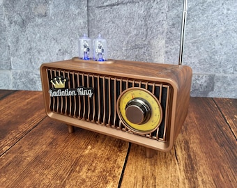 May include: A vintage-style radio with a wood-grain finish. The radio features a gold-colored grill, a tuning dial, and the text "Radiation King" with a crown logo. Two blue-lit tubes sit on top, and a silver antenna extends upwards.