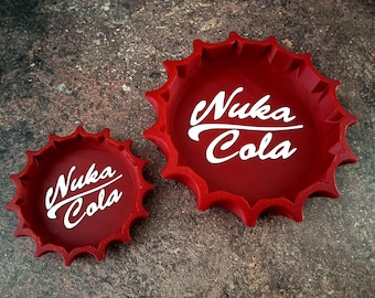 May include: Two red bottle cap-shaped trays with the text "Nuka Cola" in white script. The trays have a jagged edge design. One tray is smaller than the other.