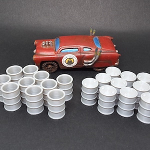 May include: A miniature, rusty red car with blue window accents and exhaust pipes, accompanied by several stacks of small, silver barrels. The car has a circular emblem on the side. The scene suggests a post-apocalyptic or industrial theme.