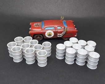 May include: A miniature, rusty red car with blue window accents and exhaust pipes, accompanied by several stacks of small, silver barrels. The car has a circular emblem on the side. The scene suggests a post-apocalyptic or industrial theme.