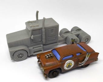 May include: A gray semi-truck and a brown vintage car are displayed against a white background. The car has blue windows and a circular emblem. The truck is a solid gray color.