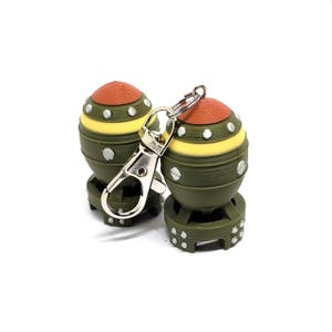 May include: Two novelty keychains shaped like miniature bombs. The keychains are olive green with yellow and orange accents. Each has a silver-colored clasp for attaching to keys or a bag. The keychains are on a white background.