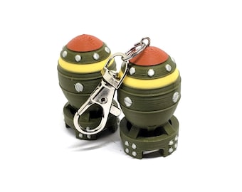 May include: Two novelty keychains shaped like miniature bombs. The keychains are olive green with yellow and orange accents. Each has a silver-colored clasp for attaching to keys or a bag. The keychains are on a white background.