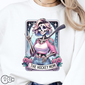 以下が含まれることがあります： ホッケーのスティックを持ったスケルトンの女性のグラフィックがプリントされた白いスウェットシャツ。グラフィックはピンク、ブルー、ホワイトのカラーで、"The Hockey Mom"というテキストが含まれています。