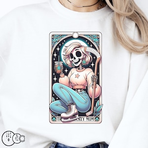 May include: White sweatshirt featuring a graphic of a skeleton woman in blue jeans holding a hockey stick. The graphic is in a vintage style with a border and the text "The Hockey Mom".