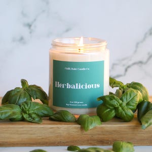 May include: A clear glass candle jar with a teal label that reads "Chill Babe Candle Co. Herbalicious 8 oz / 226 grams Soy Coconut Wax Candle". The candle is lit and surrounded by green basil leaves.