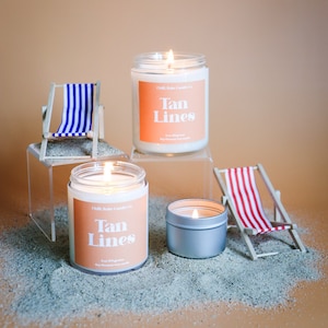 May include: Three lit candles with the text "Tan Lines" on the label, along with miniature beach chairs. One candle is in a silver tin, and the others are in glass jars. The scene is set on a sandy surface.