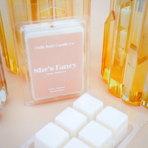 May include: A clear plastic container with six white wax melts. The container is labeled "Chill, Babe Candle Co. She's Fancy Amber - Driftwood 2.75 oz / 78 grams Soy Wax Melt"