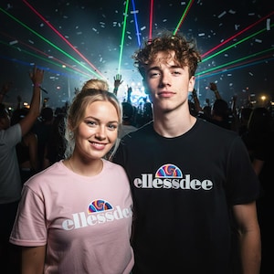 Può includere: Una donna con una maglietta rosa e un uomo con una maglietta nera, entrambi con il logo "ellessdee", posano davanti a una folla a un concerto. Lo sfondo presenta luci laser colorate e coriandoli.