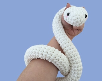 Peut inclure: Un jouet serpent en crochet blanc avec des yeux noirs et des joues roses. Le serpent est enroulé dans la main d'une personne.