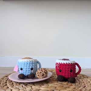 May include: Two crocheted mugs with smiling faces, one blue and one red, with chocolate chip cookies on a woven placemat.