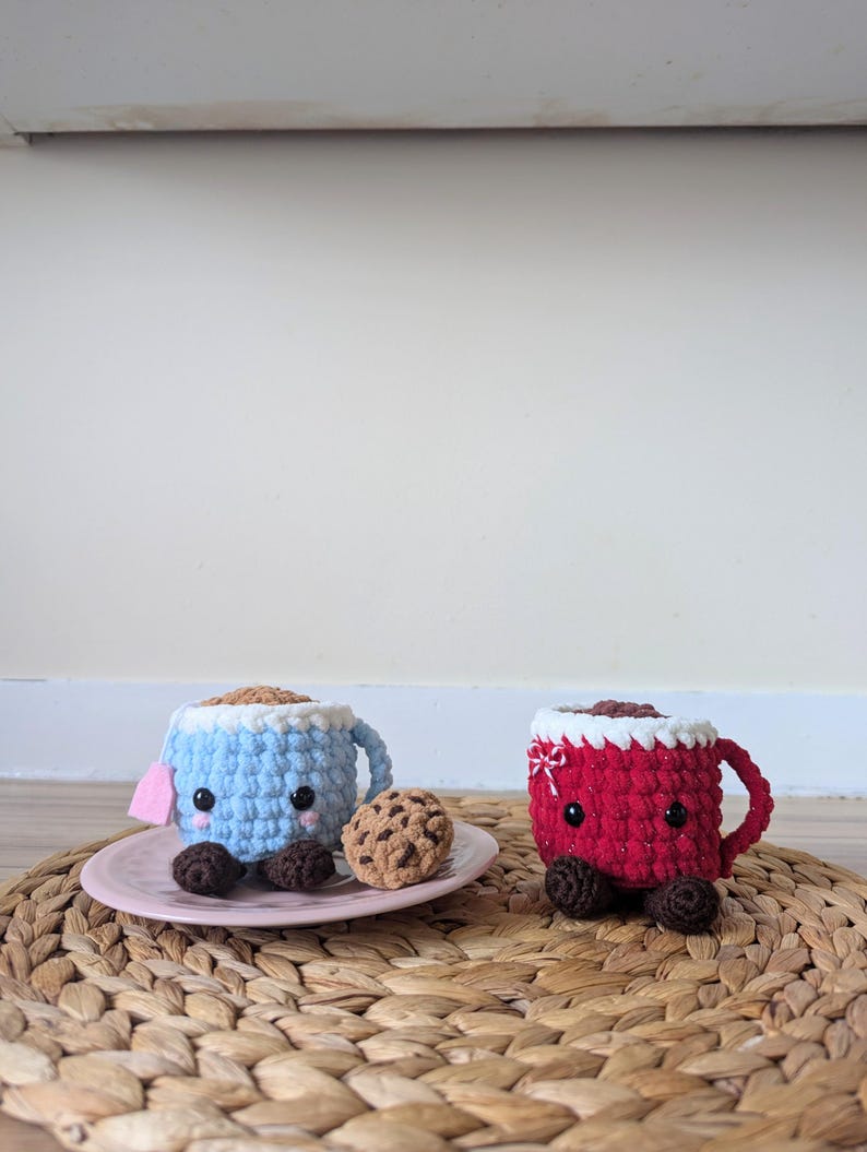 May include: Two crocheted mugs with smiling faces, one blue and one red, with chocolate chip cookies on a woven placemat.