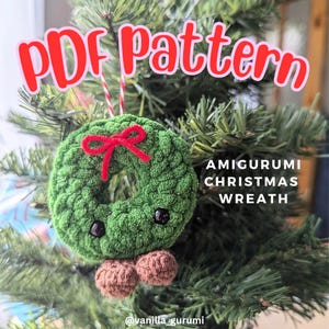 May include: A green amigurumi Christmas wreath ornament with a red bow and brown legs. The ornament is hanging on a Christmas tree. The text "PDF pattern" is in the top left corner of the image. The text "AMIGURUMI CHRISTMAS WREATH" is in the bottom right corner of the image. The text "@vanilla_gurumi" is in the bottom right corner of the image.