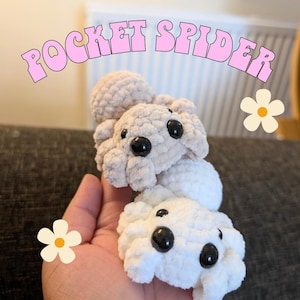 May include: Two crocheted spiders in light beige and white, with black eyes, are stacked on top of each other. The text "POCKET SPIDER" is arched above the spiders in pink. A hand holds the spiders, with a flower graphic on the side.