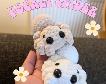 Peut inclure: Deux araignées crochetées beige clair et blanches, avec des yeux noirs, sont empilées l'une sur l'autre. Le texte "POCKET SPIDER" est arqué au-dessus des araignées en rose. Une main tient les araignées, avec un motif de fleur sur le côté.