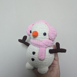 K&ouml;nnte beinhalten: Ein wei&szlig;er geh&auml;kelter Schneemann mit rosa Ohrensch&uuml;tzern und einem rosa Schal. Der Schneemann hat schwarze Augen und eine orange Nase. Der Schneemann hat zwei braune geh&auml;kelte Arme.