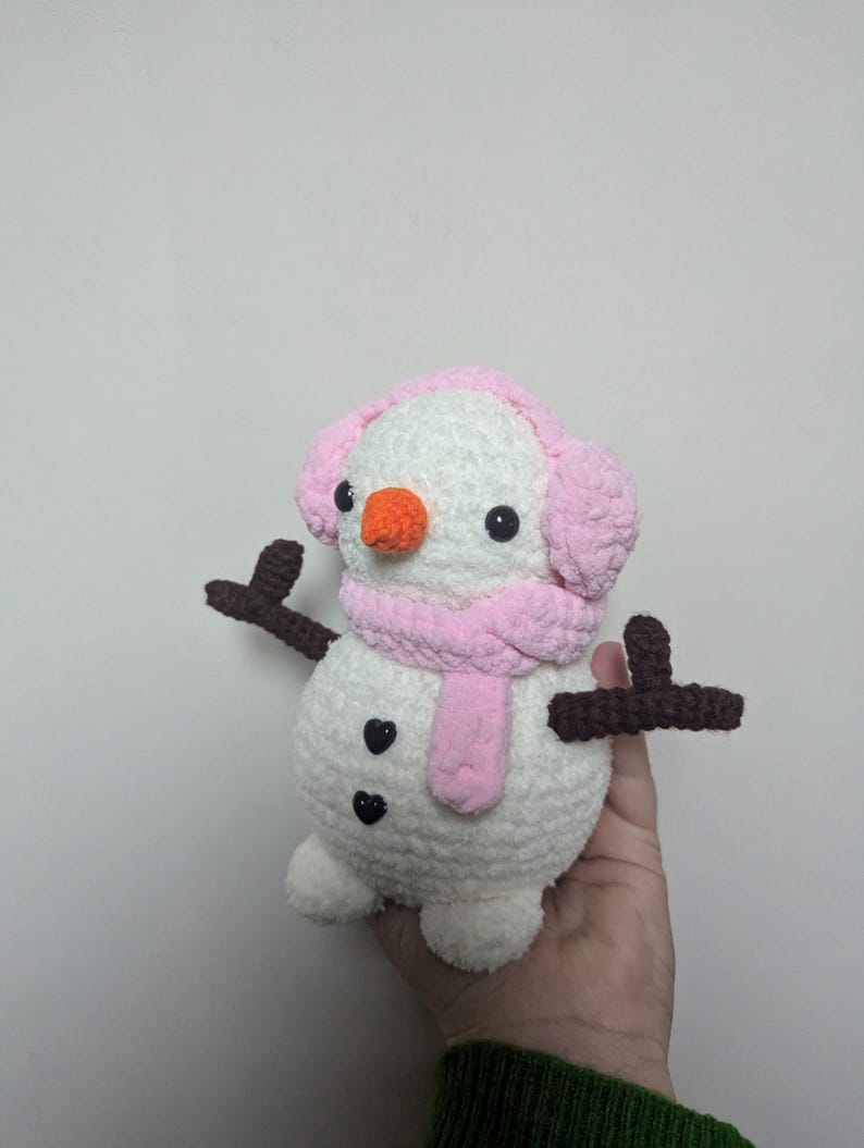 K&ouml;nnte beinhalten: Ein wei&szlig;er geh&auml;kelter Schneemann mit rosa Ohrensch&uuml;tzern und einem rosa Schal. Der Schneemann hat schwarze Augen und eine orange Nase. Der Schneemann hat zwei braune geh&auml;kelte Arme.