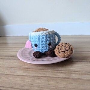 May include: A crocheted blue teacup with a pink teabag and brown eyes, sitting on a pink saucer with a crocheted brown chocolate chip cookie.