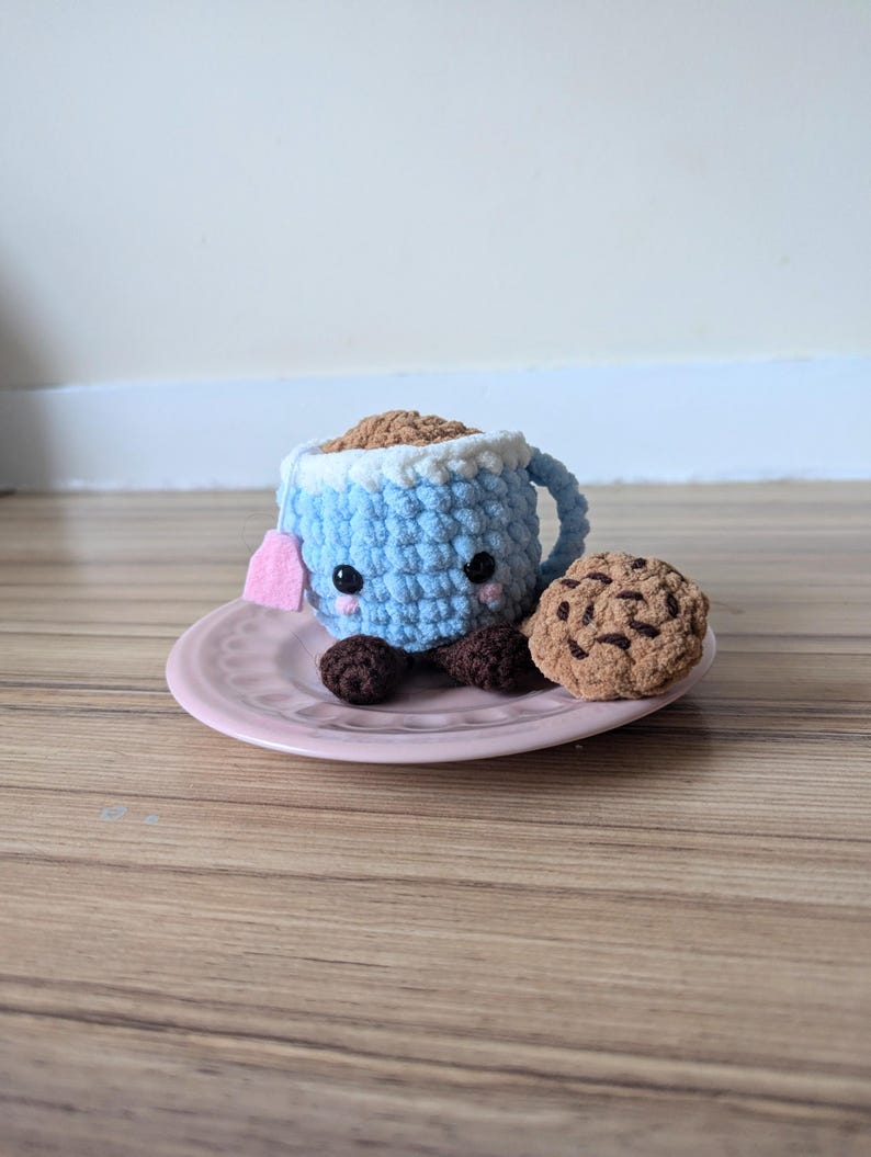 May include: A crocheted blue teacup with a pink teabag and brown eyes, sitting on a pink saucer with a crocheted brown chocolate chip cookie.