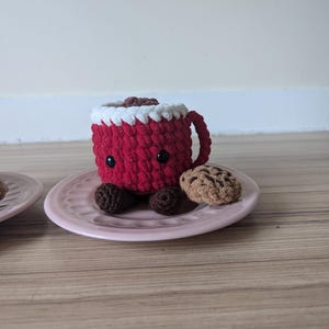 May include: A red crocheted mug with white frosting and chocolate chips, sitting on a pink plate with a brown crocheted cookie.