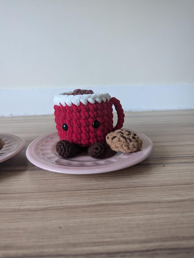 May include: A red crocheted mug with white frosting and chocolate chips, sitting on a pink plate with a brown crocheted cookie.