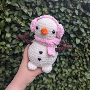 K&ouml;nnte beinhalten: Ein wei&szlig;er geh&auml;kelter Schneemann mit rosa Ohrensch&uuml;tzern und einem Schal. Der Schneemann hat schwarze Augen, eine orange Nase und zwei schwarze Kn&ouml;pfe.