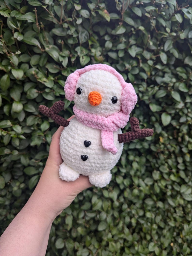 K&ouml;nnte beinhalten: Ein wei&szlig;er geh&auml;kelter Schneemann mit rosa Ohrensch&uuml;tzern und einem Schal. Der Schneemann hat schwarze Augen, eine orange Nase und zwei schwarze Kn&ouml;pfe.