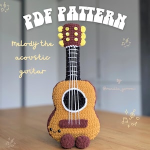 May include: A crochet acoustic guitar toy with a brown body, black neck, and yellow tuning pegs. The text reads PDF PATTERN, Melody the acoustic guitar, and By @vanilla_gurumi. The guitar features white yarn strings, a black sound hole, and a small embroidered face.