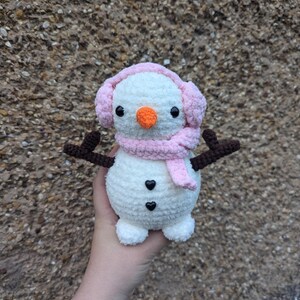 K&ouml;nnte beinhalten: Ein wei&szlig;er geh&auml;kelter Schneemann mit einem rosa Schal und Ohrensch&uuml;tzern. Der Schneemann hat schwarze Augen, eine orange Nase und zwei schwarze herzf&ouml;rmige Kn&ouml;pfe.