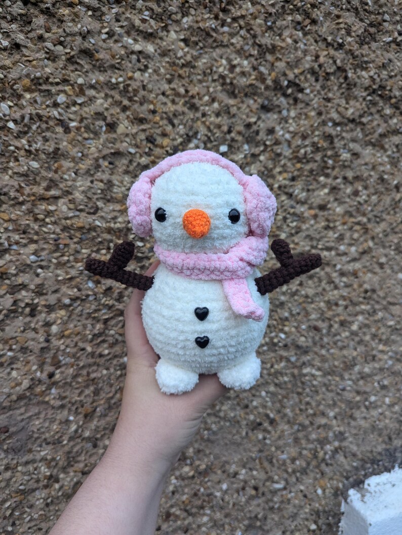 K&ouml;nnte beinhalten: Ein wei&szlig;er geh&auml;kelter Schneemann mit einem rosa Schal und Ohrensch&uuml;tzern. Der Schneemann hat schwarze Augen, eine orange Nase und zwei schwarze herzf&ouml;rmige Kn&ouml;pfe.