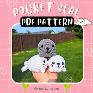 May include: Three crocheted seal plushies, one grey and two white, are stacked on top of each other. The plushies are small and have cute faces. The image is a close-up of the plushies, and the background is a blurry green lawn. The text "POCKET SEAL PDF PATTERN" is at the top of the image.