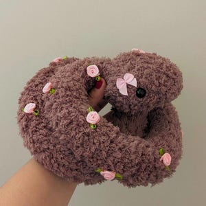 May include: A brown teddy bear toy with a pink bow and small pink rose decorations. The bear has a soft, fluffy texture and a circular shape. The pink roses are attached to green stems.