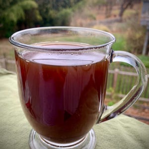May include: A clear glass mug filled with a dark brown liquid, possibly tea or coffee. The mug has a handle and sits on a small, clear base. The background is blurred, showing greenery and a wooden fence.