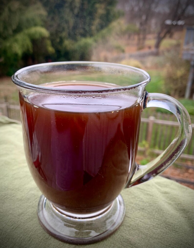 May include: A clear glass mug filled with a dark brown liquid, possibly tea or coffee. The mug has a handle and sits on a small, clear base. The background is blurred, showing greenery and a wooden fence.