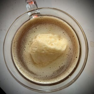 May include: A clear glass mug filled with a dark brown drink, with a large scoop of pale yellow ice cream floating on top. The drink has a foamy texture with bubbles. The mug's handle is visible.
