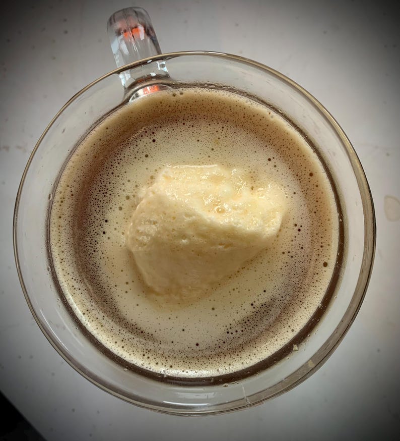 May include: A clear glass mug filled with a dark brown drink, with a large scoop of pale yellow ice cream floating on top. The drink has a foamy texture with bubbles. The mug's handle is visible.
