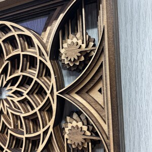 Puede incluir: Una pieza de arte mural de madera en capas con un dise&ntilde;o geom&eacute;trico. La obra de arte presenta un patr&oacute;n circular y acentos florales, todo elaborado con madera marr&oacute;n clara y oscura. La pieza est&aacute; enmarcada y tiene un efecto tridimensional.
