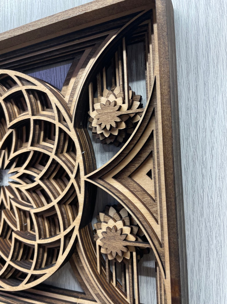 Puede incluir: Una pieza de arte mural de madera en capas con un dise&ntilde;o geom&eacute;trico. La obra de arte presenta un patr&oacute;n circular y acentos florales, todo elaborado con madera marr&oacute;n clara y oscura. La pieza est&aacute; enmarcada y tiene un efecto tridimensional.