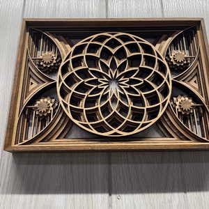 Puede incluir: Una pieza de arte mural de madera en capas con un dise&ntilde;o floral central y patrones geom&eacute;tricos. La obra de arte est&aacute; enmarcada en madera marr&oacute;n oscuro y tiene un aspecto de veta natural. El dise&ntilde;o incorpora m&uacute;ltiples capas, creando un efecto tridimensional.