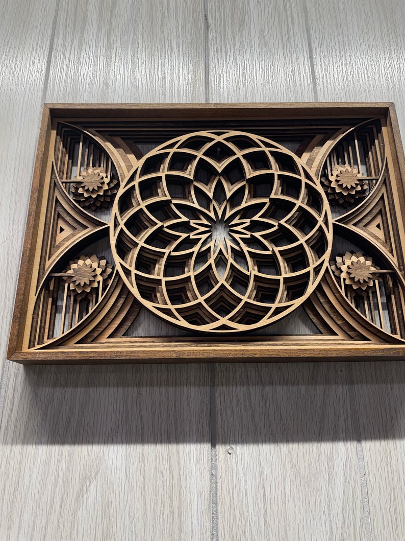 Puede incluir: Una pieza de arte mural de madera en capas con un dise&ntilde;o floral central y patrones geom&eacute;tricos. La obra de arte est&aacute; enmarcada en madera marr&oacute;n oscuro y tiene un aspecto de veta natural. El dise&ntilde;o incorpora m&uacute;ltiples capas, creando un efecto tridimensional.
