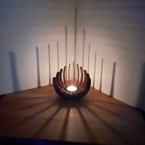 K&ouml;nnte beinhalten: Ein Kerzenhalter aus Holz mit geometrischem Design, der einen Schatten auf eine Holzoberfl&auml;che wirft. Der Kerzenhalter ist angez&uuml;ndet und die Kerze ist wei&szlig;.